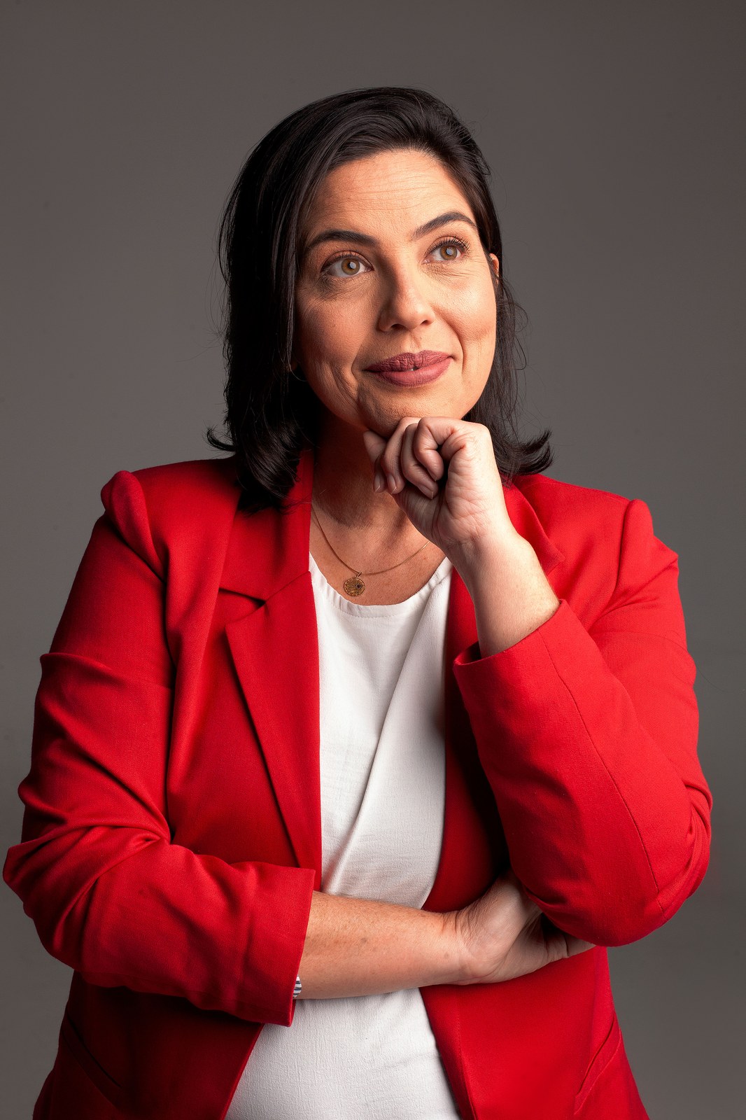 Juliana Fraga, de blazer vermelho, em retrato de campanha, com a mão apoiada no rosto.